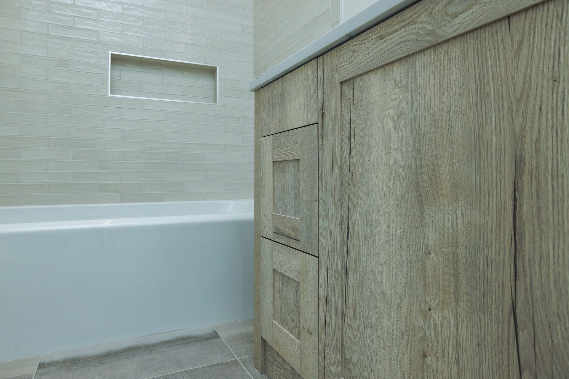A side view of a bathroom vanity with light wood cabinets, positioned next to a white bathtub with a tiled wall niche.