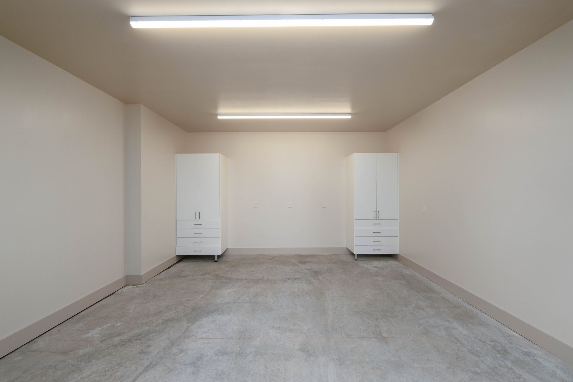 A minimalist, brightly lit room with beige walls, a grey concrete floor, and two white cabinets against the back wall.