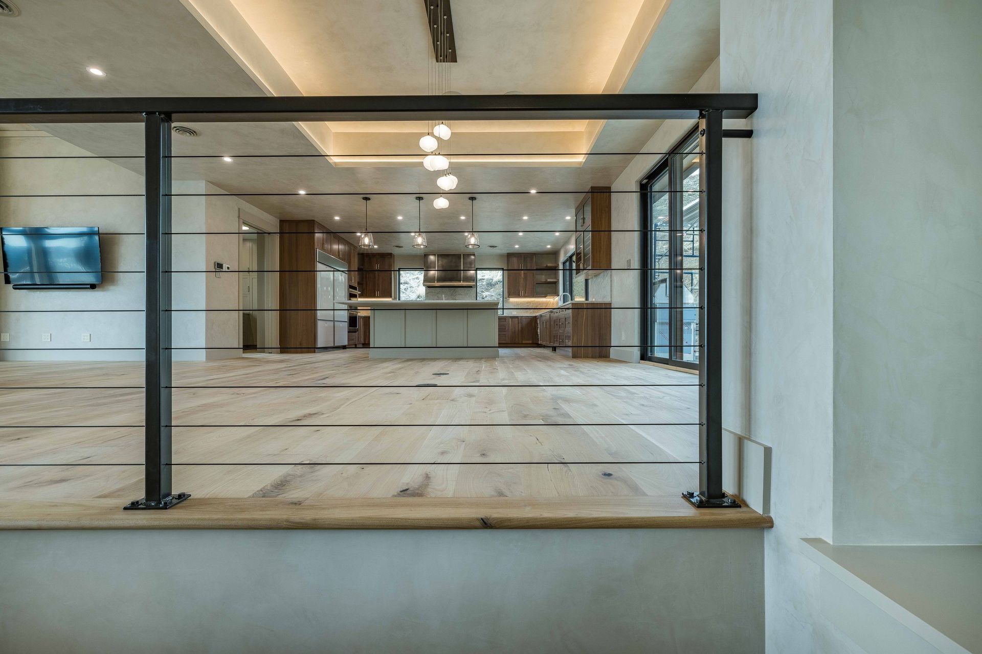 A cable railing overlooks an open-concept kitchen with wooden cabinetry, a central island, and light hardwood floors.