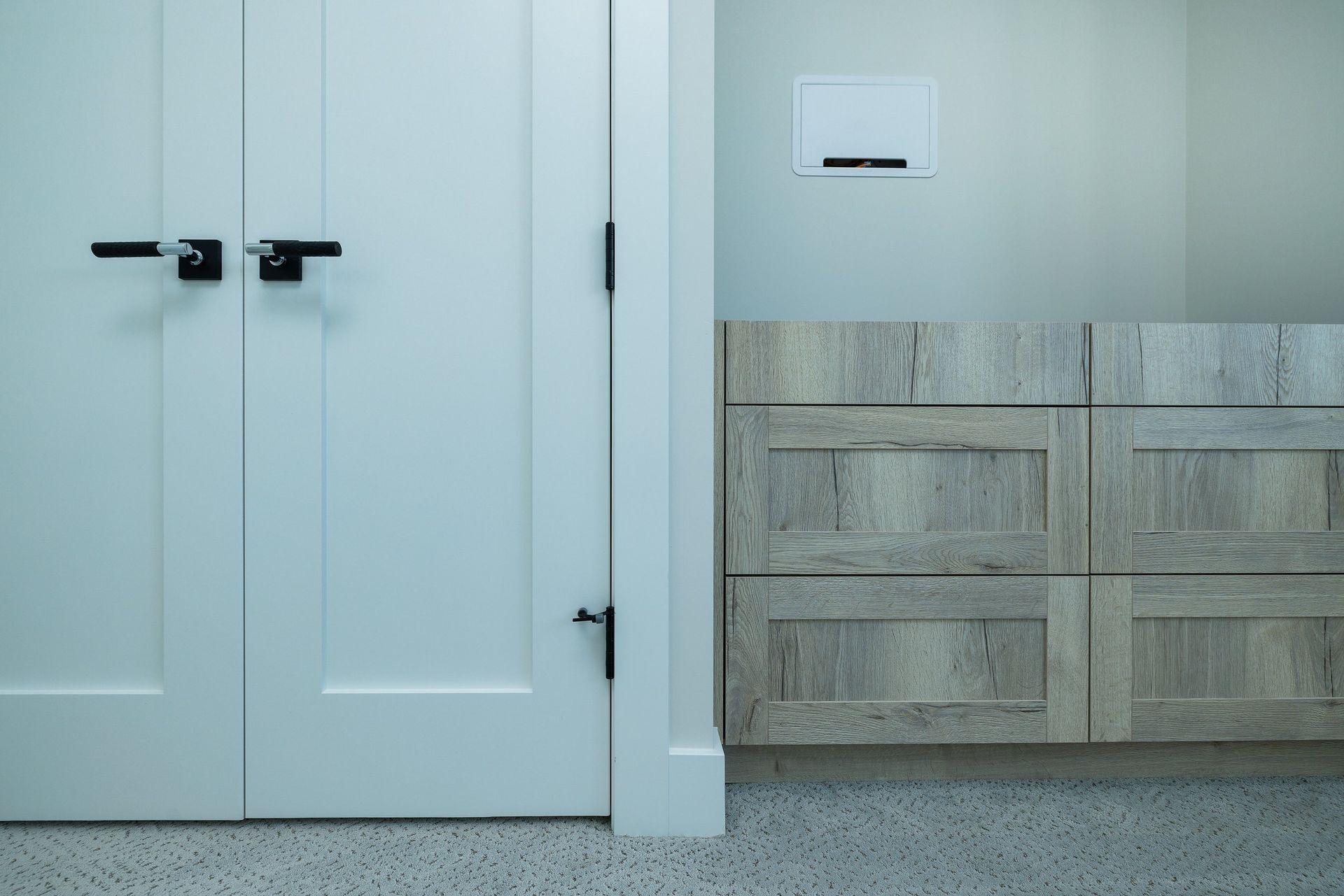White double doors with black hardware stand next to a light wood cabinet with a white wall-mounted toilet control panel.