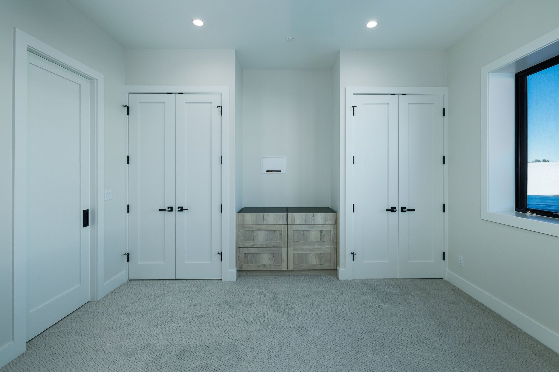 A room with white walls, gray carpet, three white doors, and a small built-in wooden cabinet centered on the back wall.