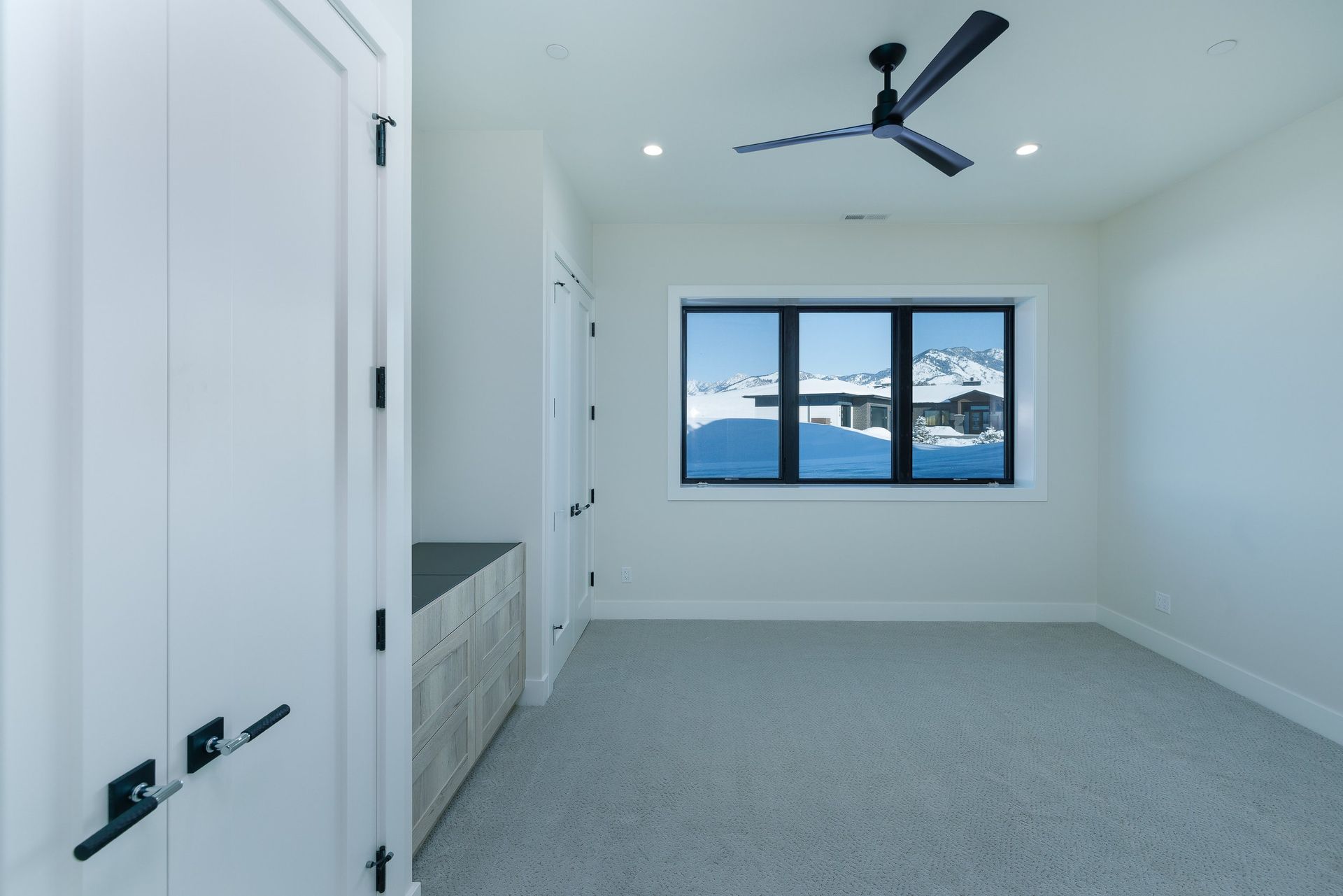 A bright, empty bedroom with white walls, grey carpeting, a black ceiling fan, and a window overlooking a snowy landscape.