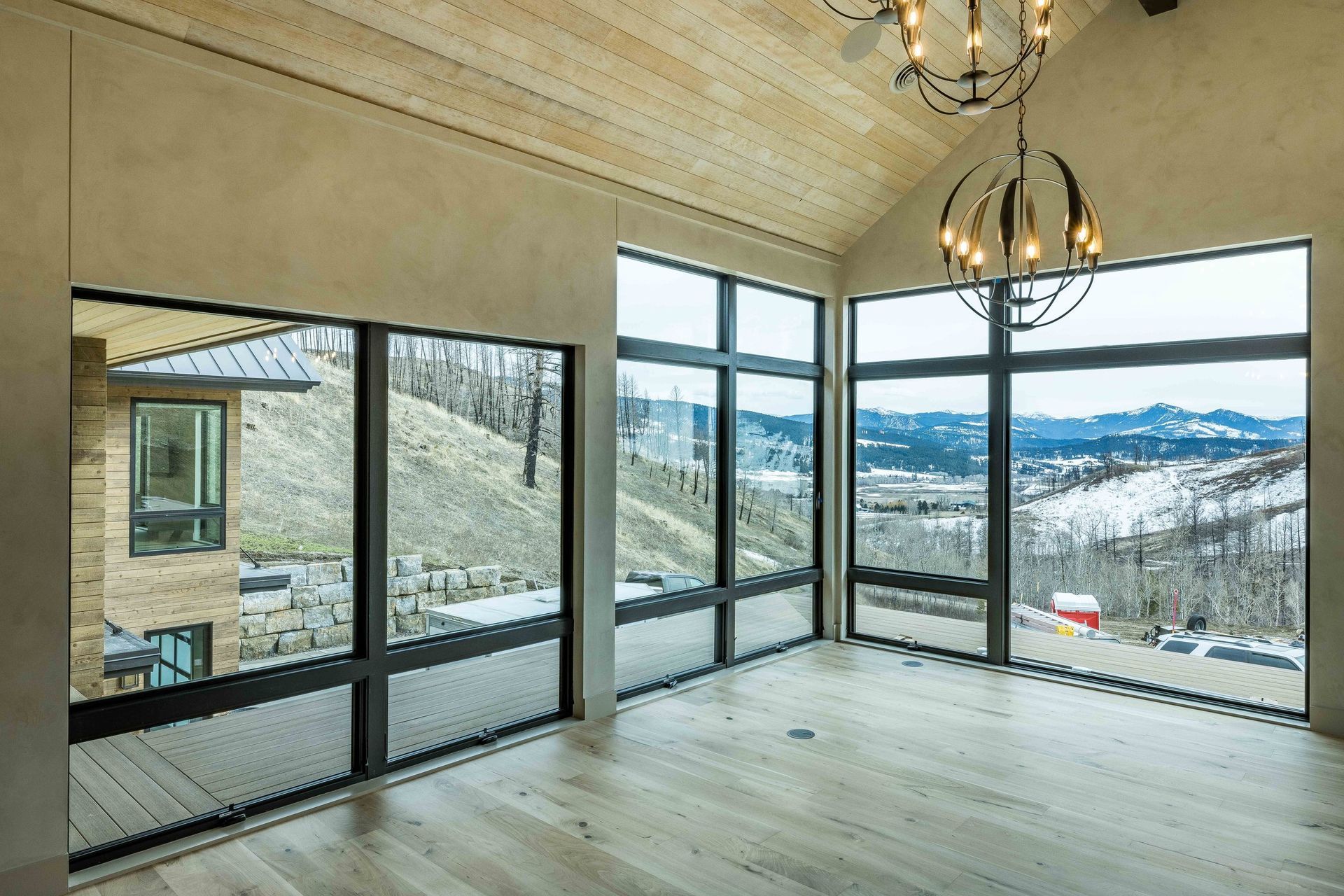 A sunlit room with light wood floors, a vaulted wood ceiling, and floor-to-ceiling windows showing a mountain view.