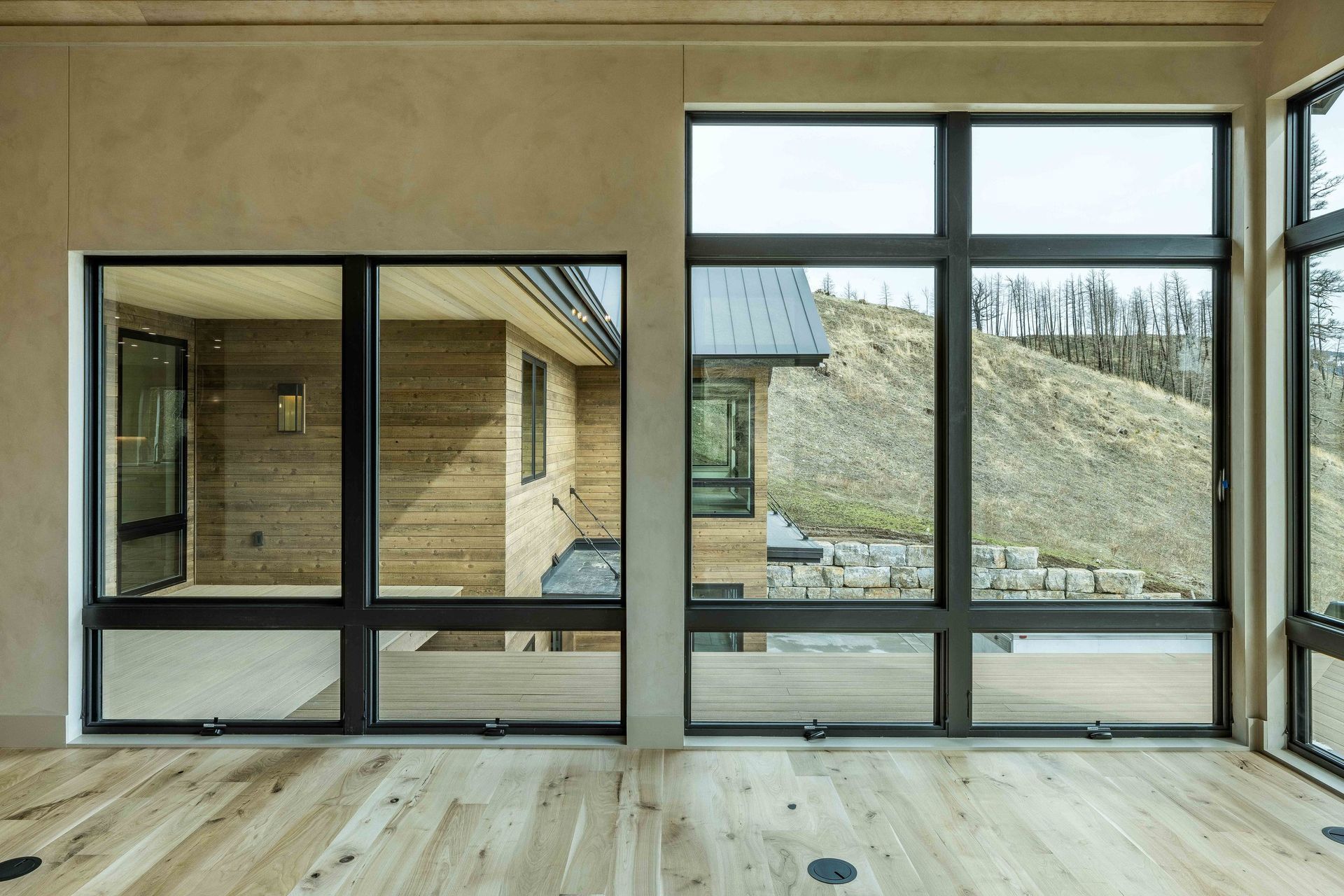A view through large, black-framed windows looking out onto a wooden deck and a hillside.