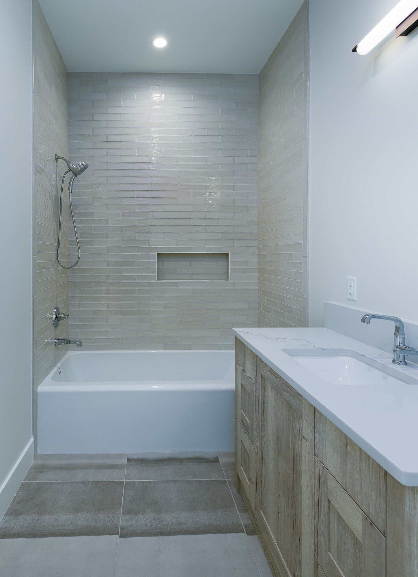 A modern bathroom featuring a white bathtub, gray tiled shower walls, a built-in niche, and a wooden vanity with a sink.