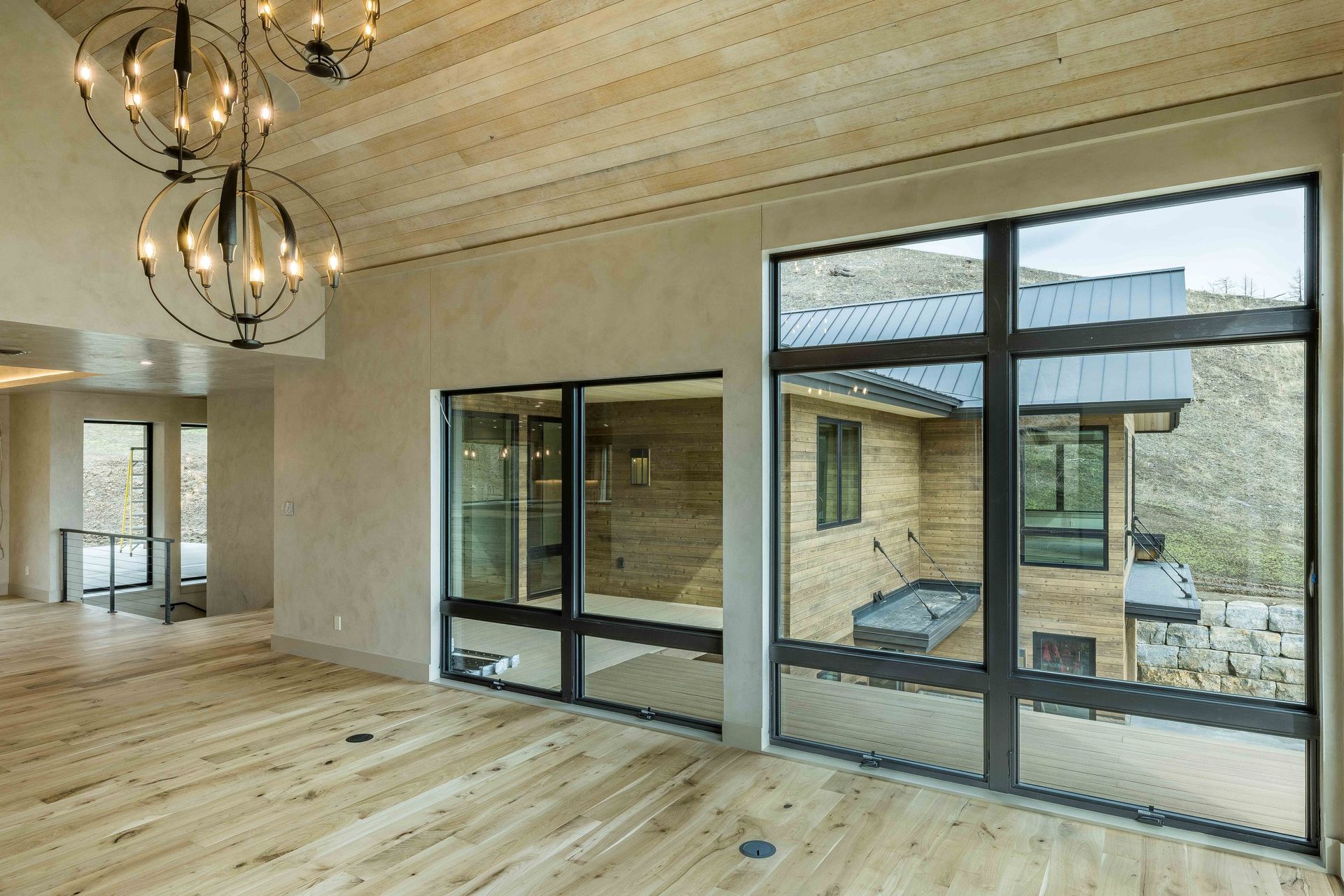 A modern room with light wood floors, a vaulted wood-paneled ceiling, two orb chandeliers, and large floor-to-ceiling windows.