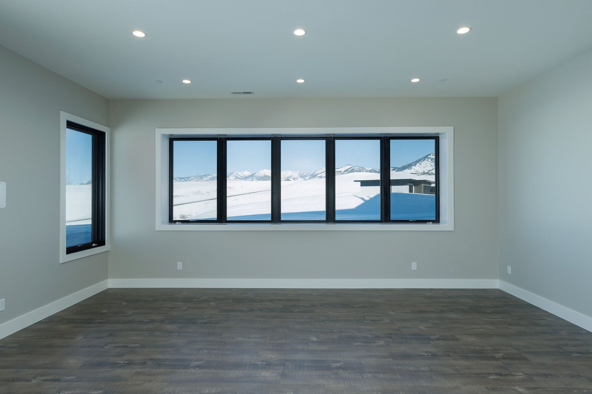 Empty room with gray walls, dark wood floors, recessed ceiling lights, and large windows overlooking a snowy landscape.