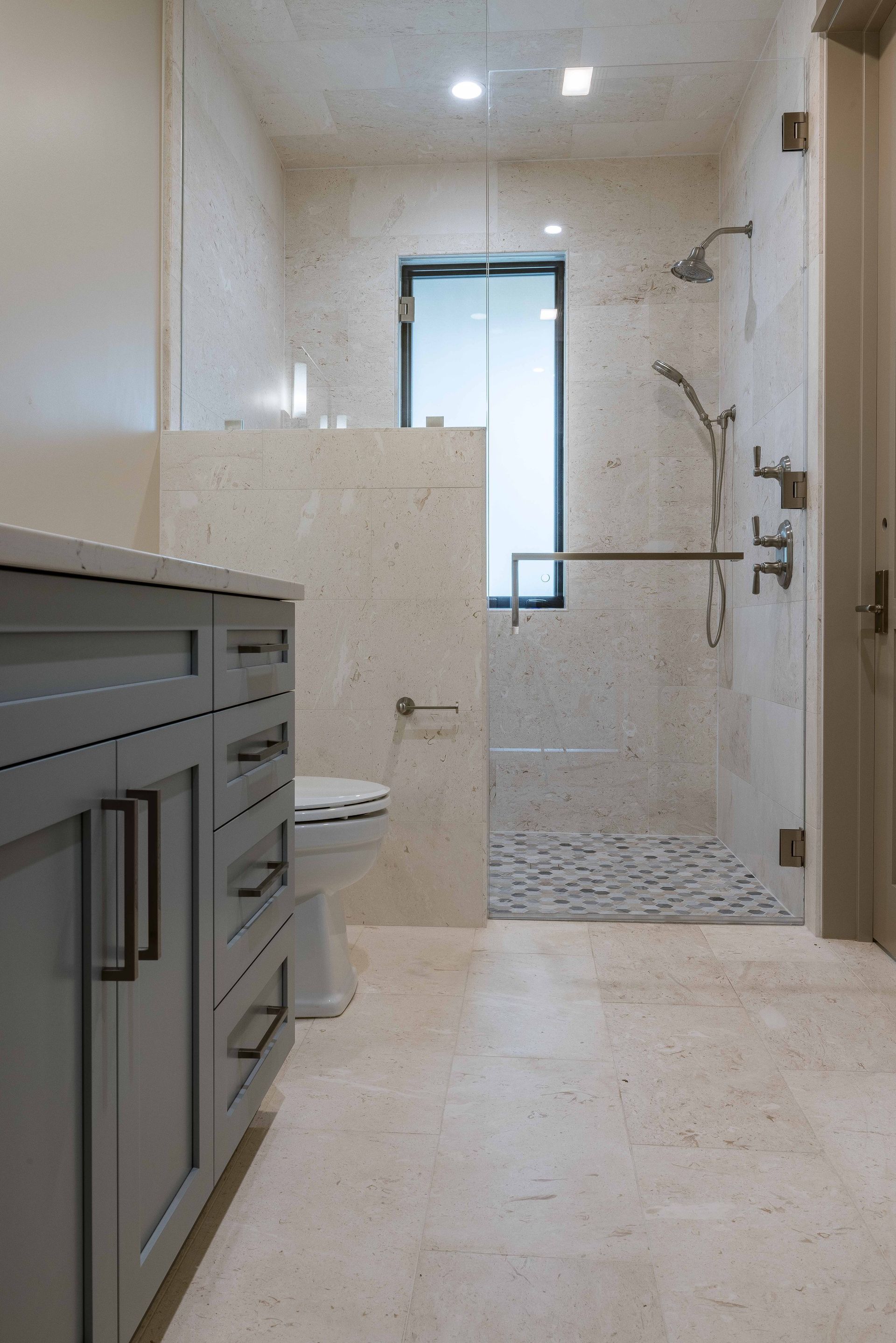 A bathroom featuring a grey vanity, a toilet, and a glass-enclosed shower with beige tile walls and patterned floor tiles.