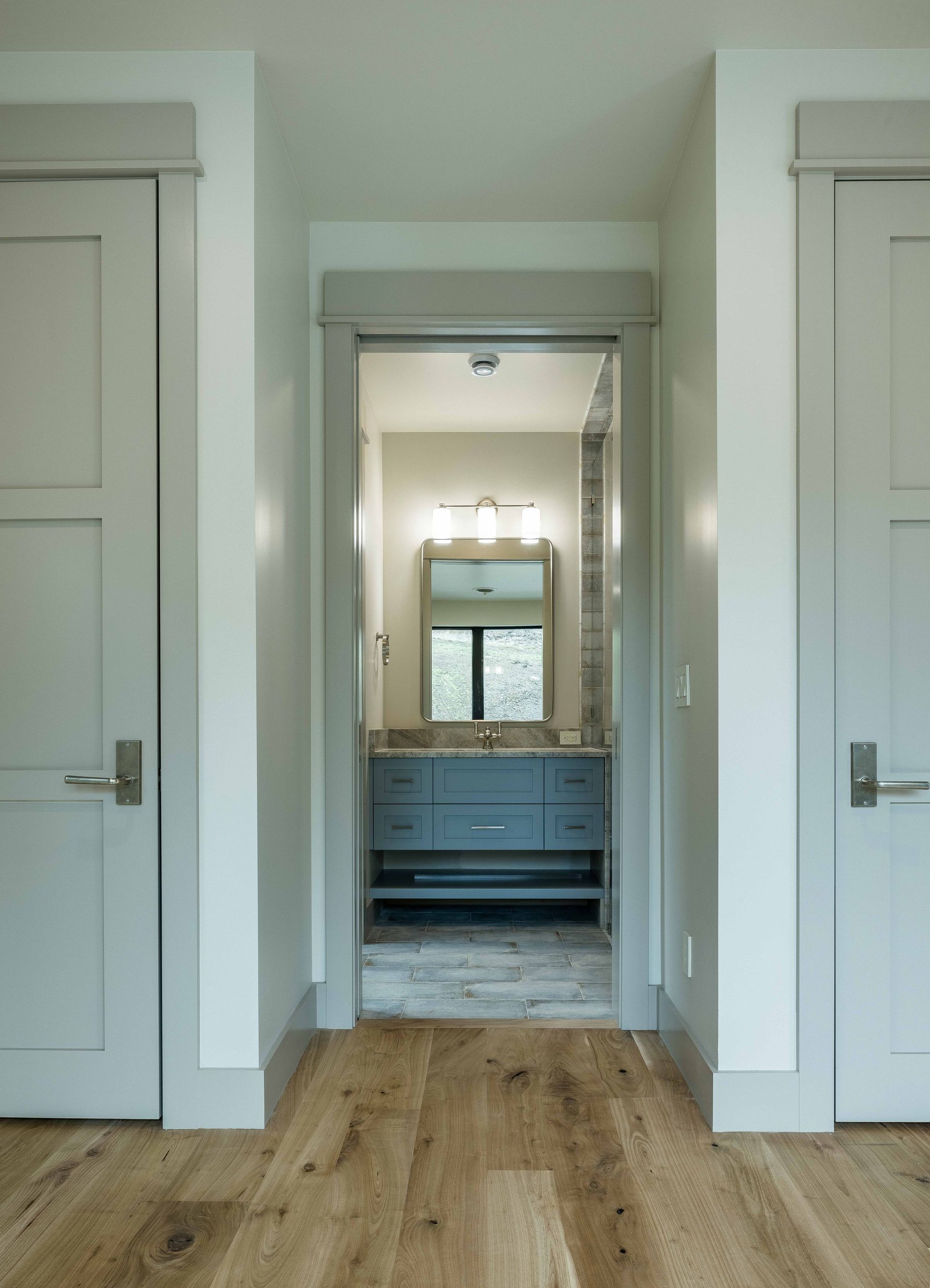 A view through a hallway with light-colored doors on either side, leading to a bathroom with a grey vanity and mirror.
