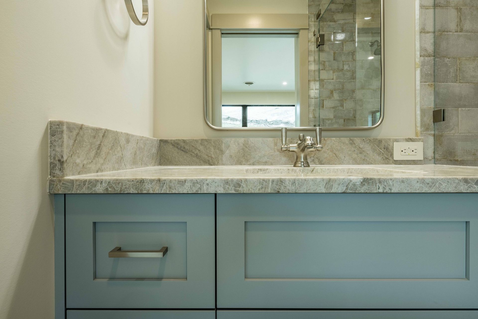 A blue bathroom vanity with a granite countertop, sink, faucet, and mirror reflecting a tiled shower.