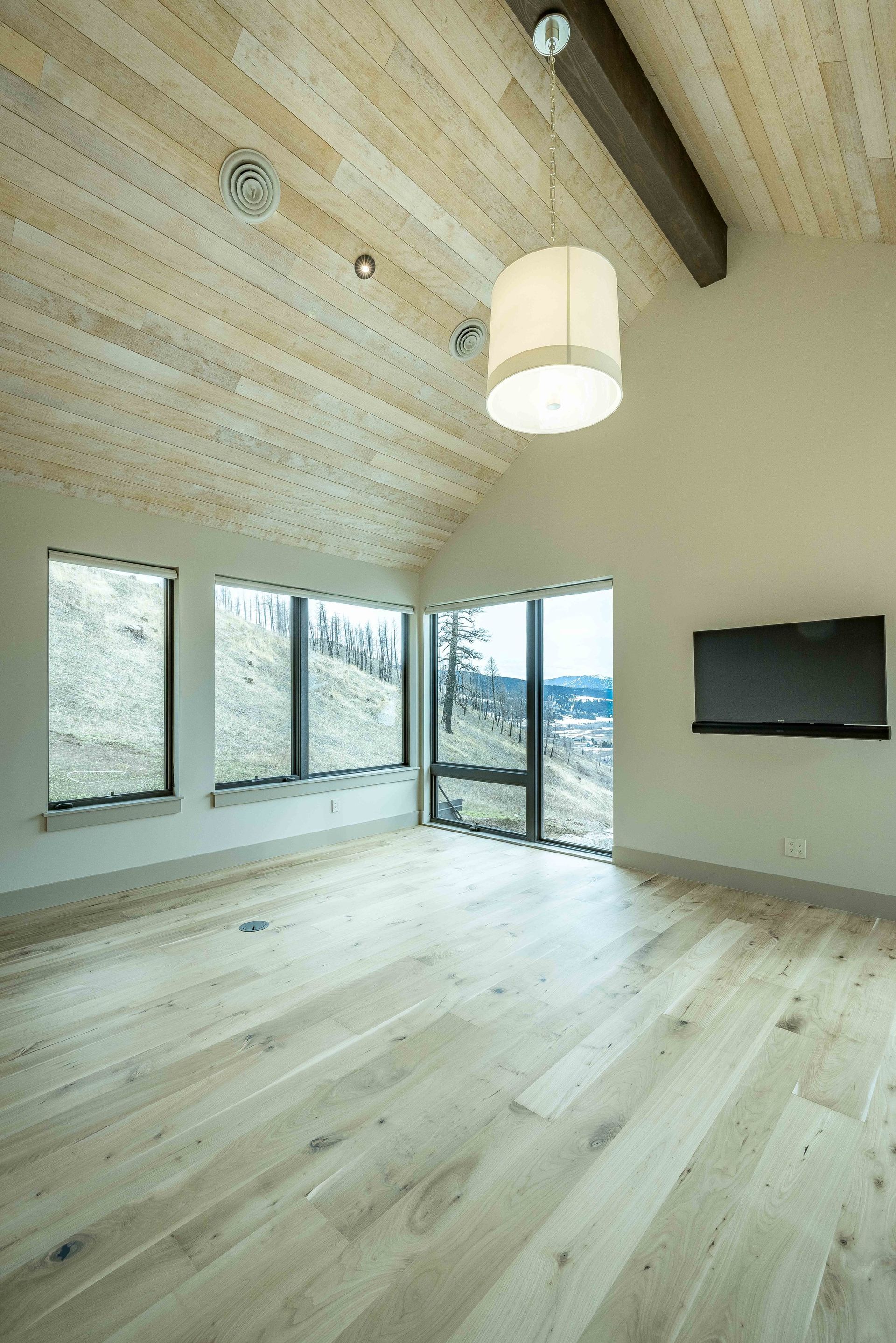 An unfurnished room with light wood floors, a vaulted ceiling, large windows with a landscape view, and a wall-mounted TV.