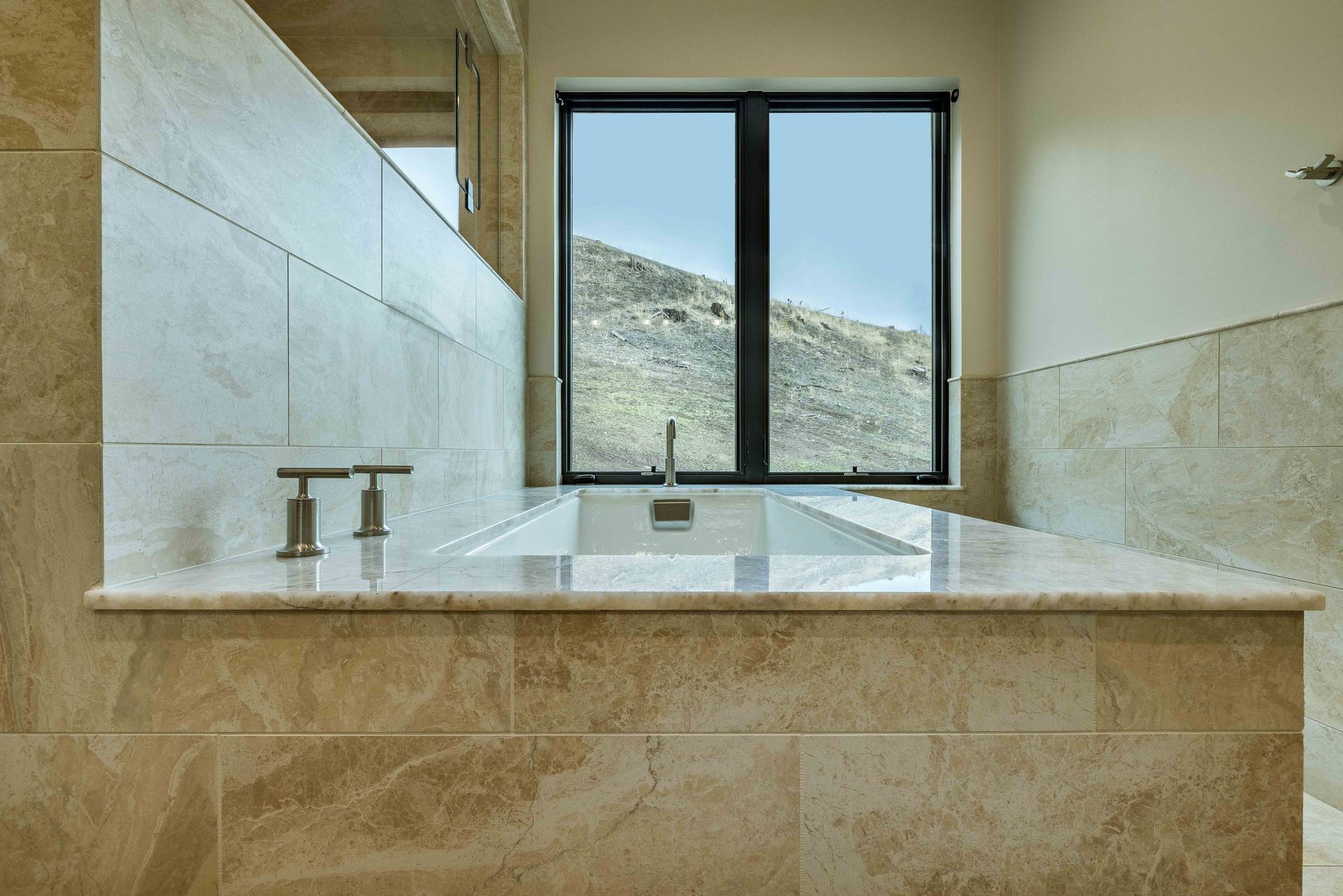 A beige tiled bathtub with metallic faucets sits beneath a window overlooking a rocky hill.