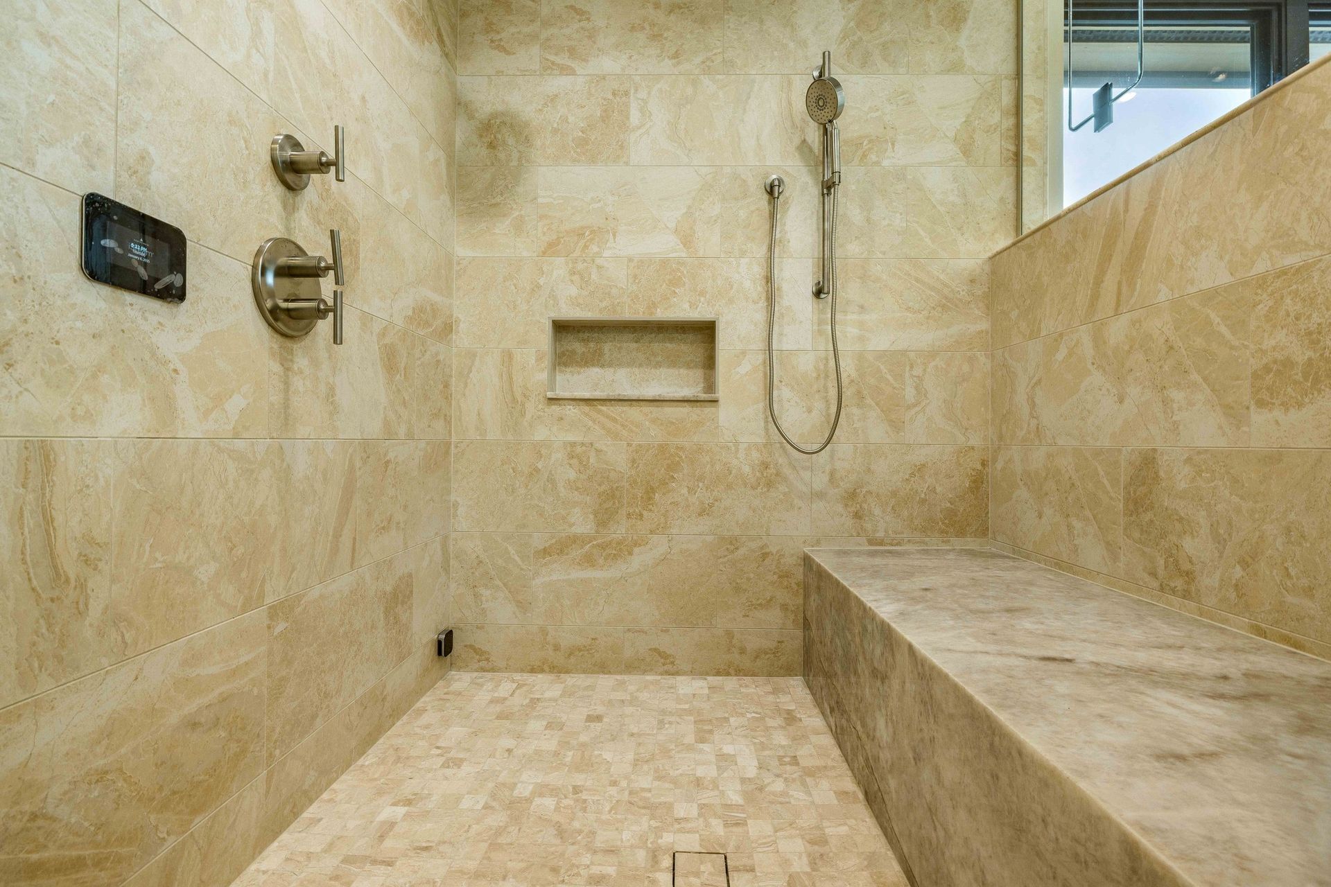 A modern, beige tiled walk-in shower with a built-in stone bench, handheld showerhead, and wall-mounted controls.
