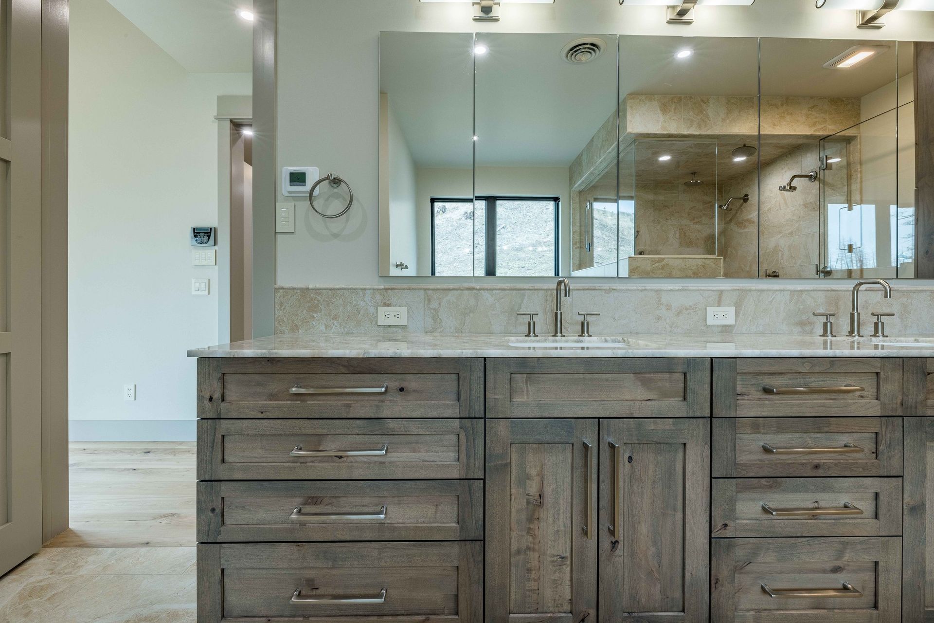 A double vanity with weathered wood cabinets, marble countertop, and large wall-to-wall mirrors in a bright bathroom.