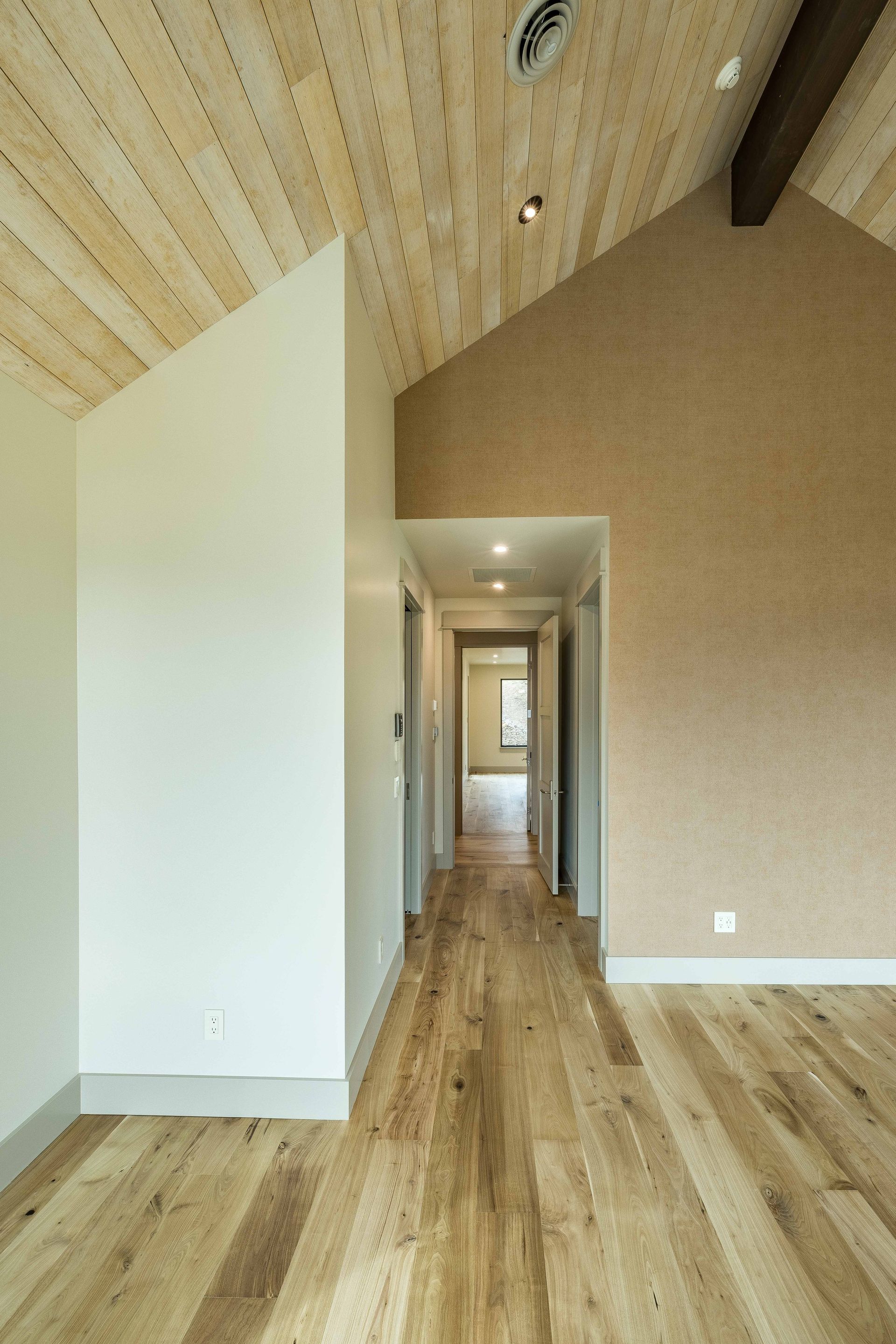 A hallway with light wood floors leads into a bright room, set against white walls and a vaulted, wood-paneled ceiling.