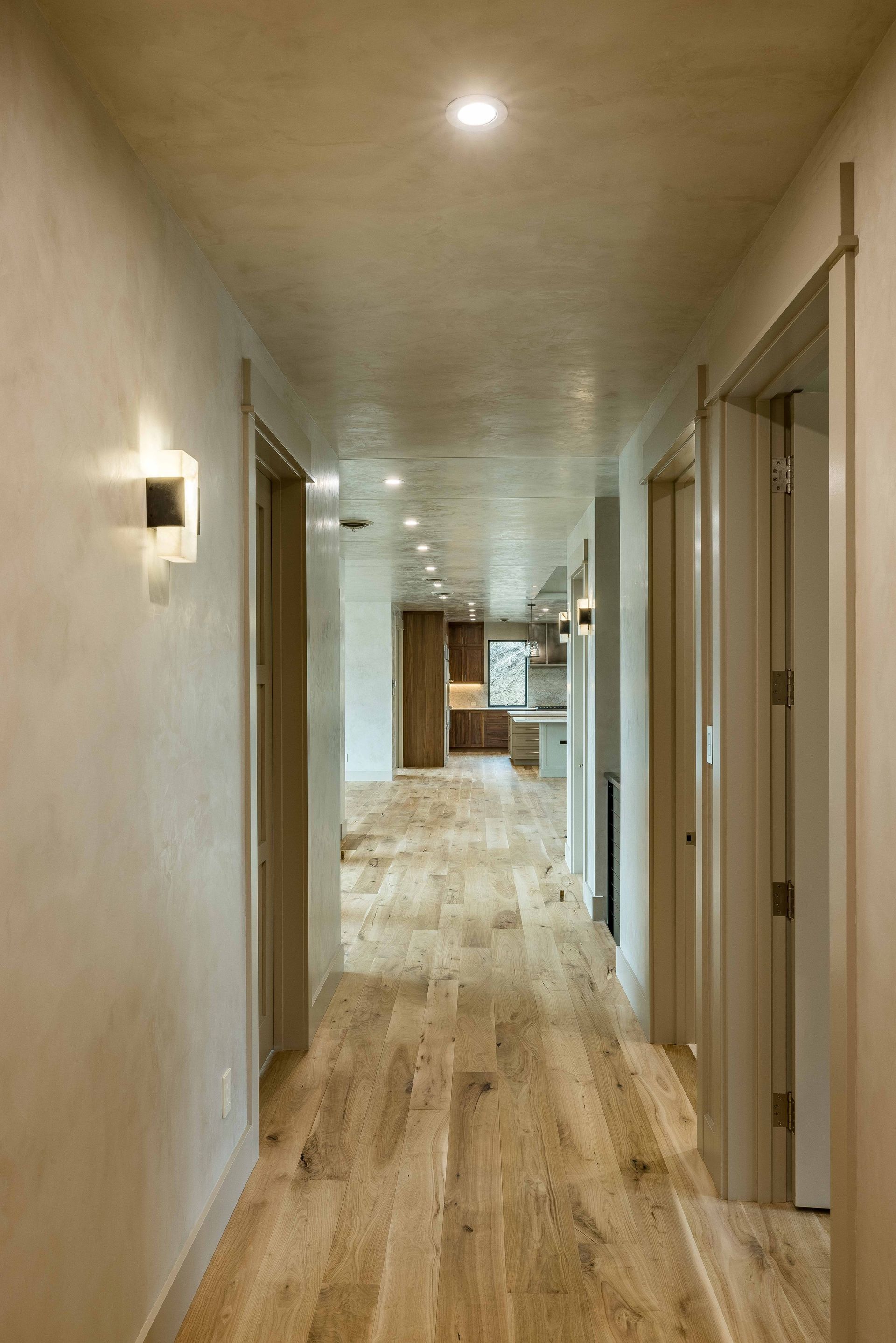 A long hallway with light wood floors and soft wall lighting, leading to a glimpse of a kitchen in the distance.