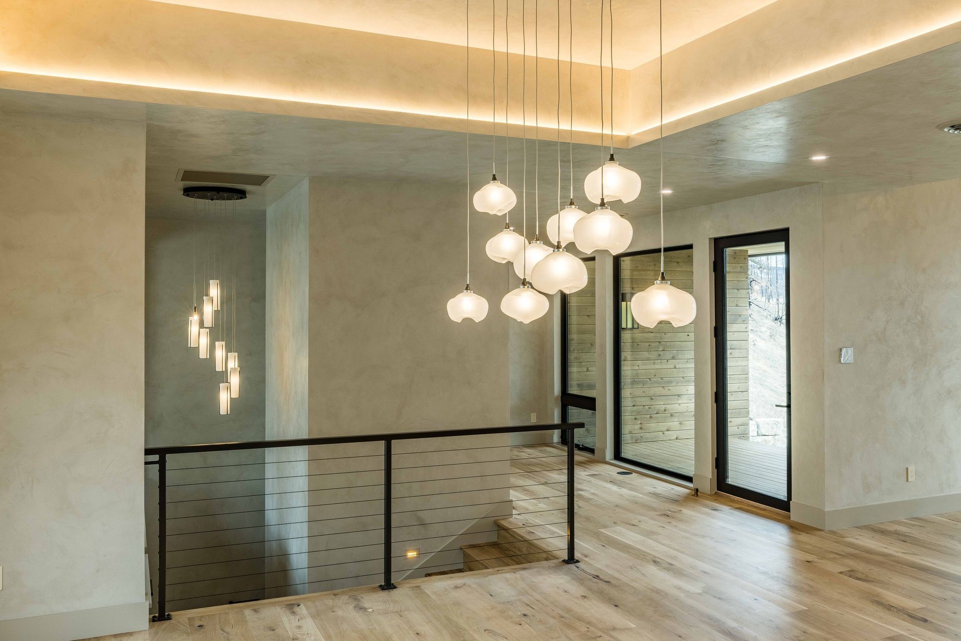 Modern foyer with light wood floors, a dark railing overlooking stairs, and two clusters of glowing white pendant lights.