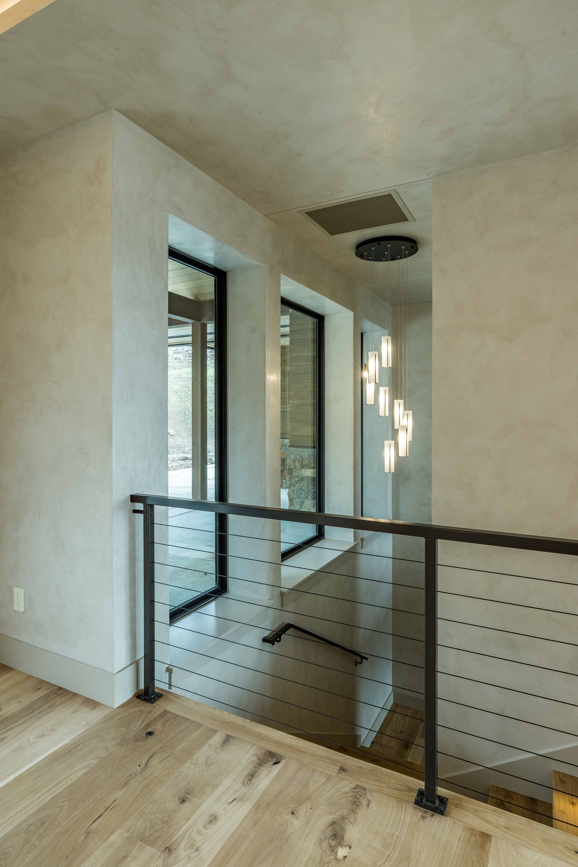 A view from a wooden floor landing with a black cable railing overlooking a stairwell with tall windows and a chandelier.