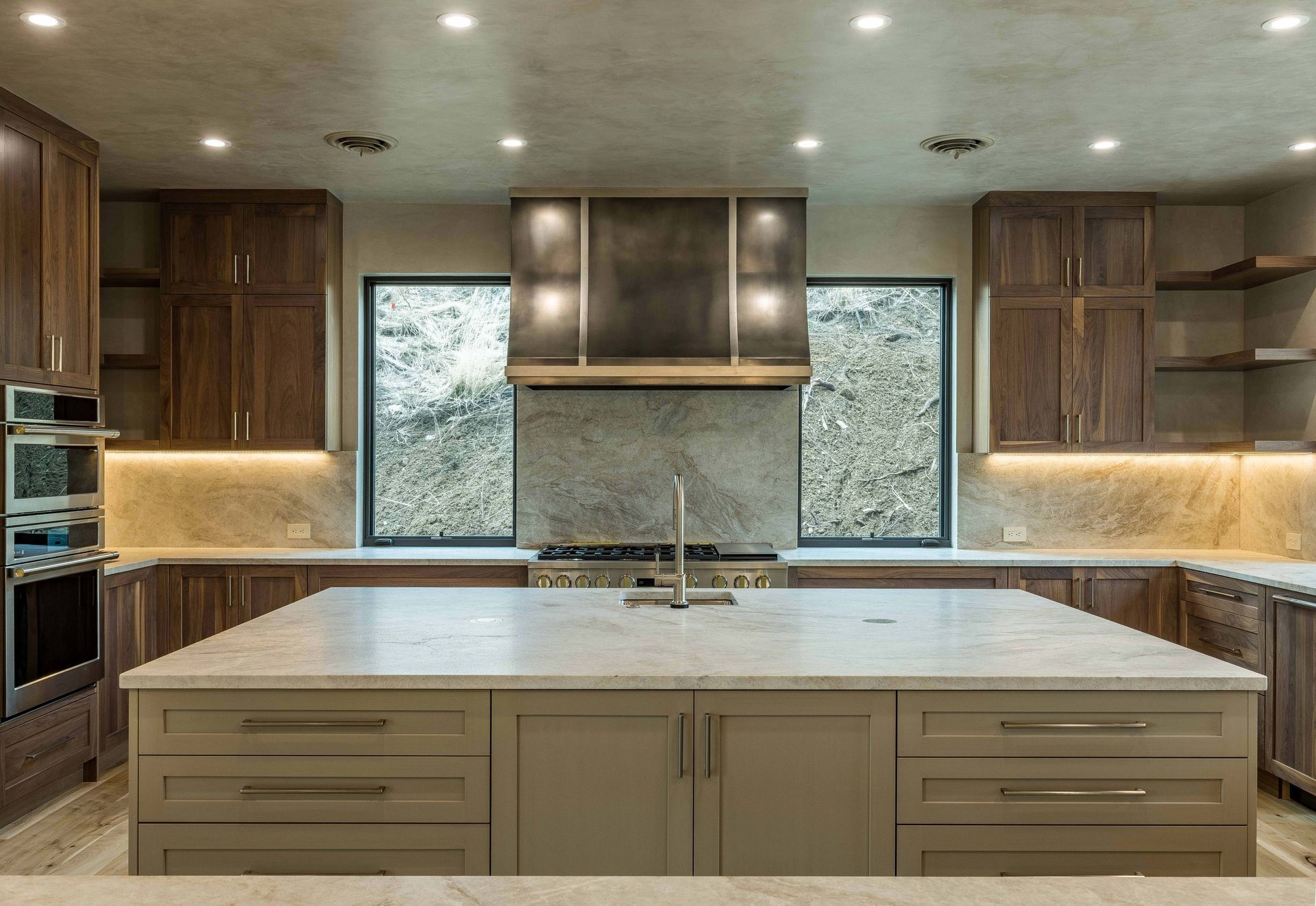 Modern kitchen with dark wood cabinetry, a large marble island, stainless appliances, and windows flanking the range hood.