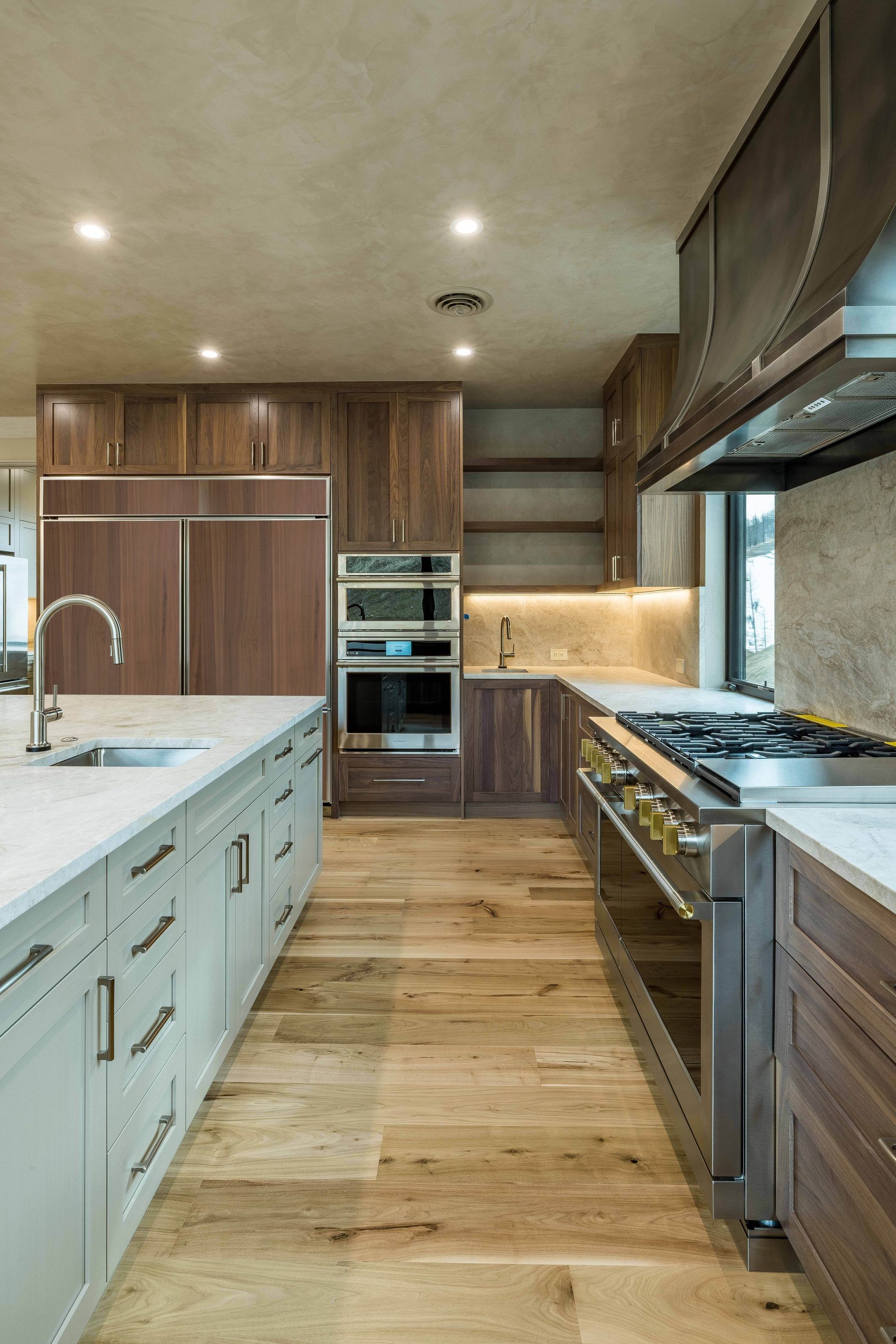 A kitchen features a long white-topped island, wooden cabinetry, stainless steel appliances, and light wood flooring.