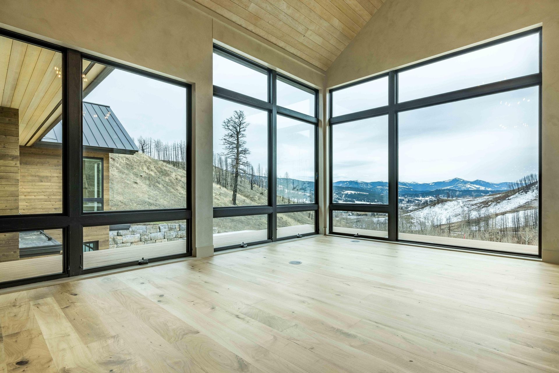 An empty room with light wood floors, a vaulted wood ceiling, and large windows revealing a snowy mountain landscape.