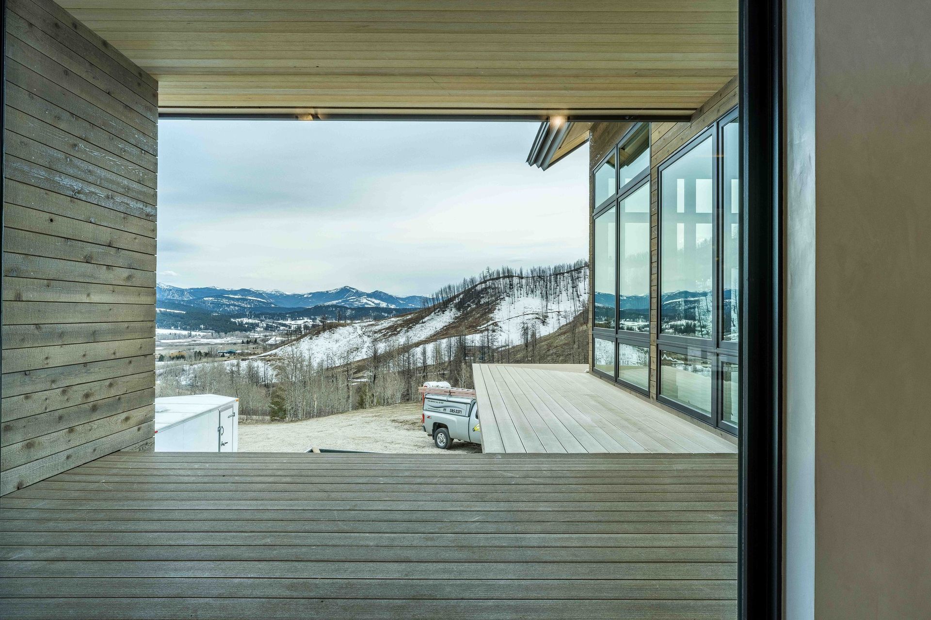 A wooden deck overlooks a snowy, mountainous landscape through a modern home's expansive, open-walled living space.