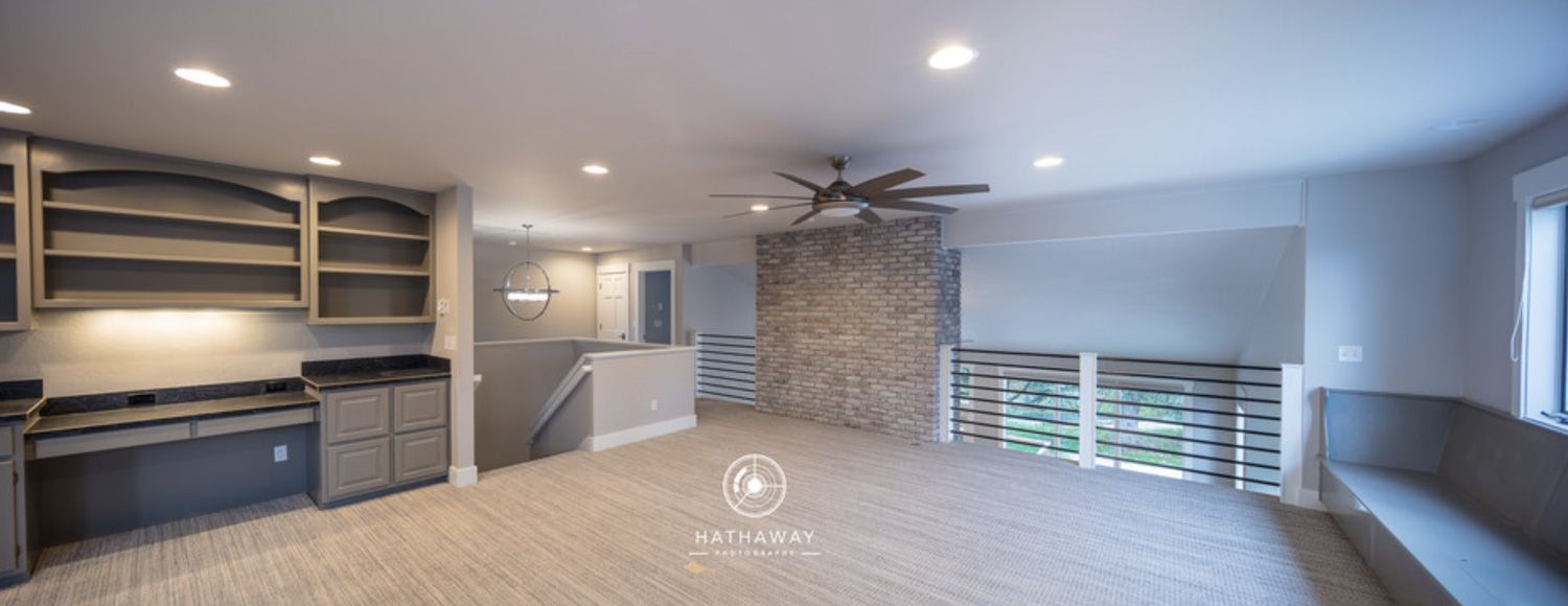 A wide-angle view of a modern room with gray built-in desks, a stone feature wall, a ceiling fan, and a loft railing.