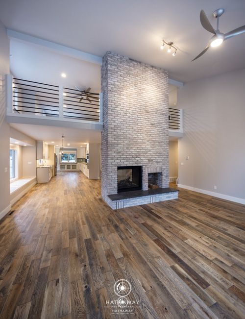 A spacious living room with hardwood floors, a large central stone fireplace, and a second-story loft with railings.