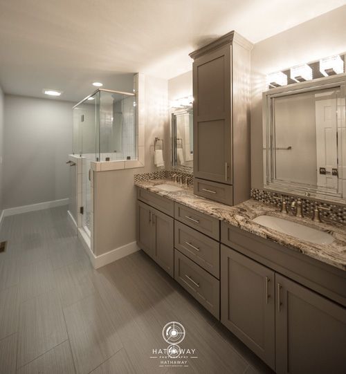 A modern bathroom with grey cabinetry, stone countertops, a tall storage tower, and a glass-enclosed shower.