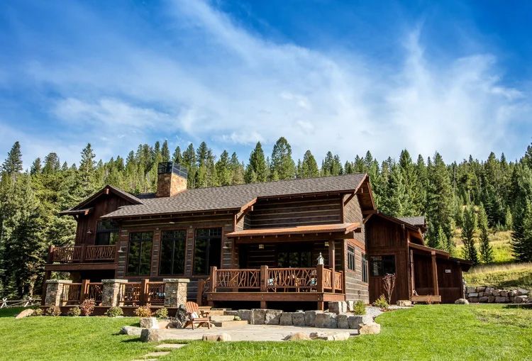 A multi-story rustic log cabin with a stone patio, wooden porch, and fireplace chimney, set against a forested hillside.