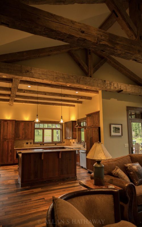 Rustic kitchen with dark wooden cabinetry, an island, and exposed timber ceiling beams, viewed from a cozy living area.