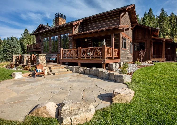 A log cabin house with a wood deck, stone patio, and large boulder accents, situated in a forested landscape.