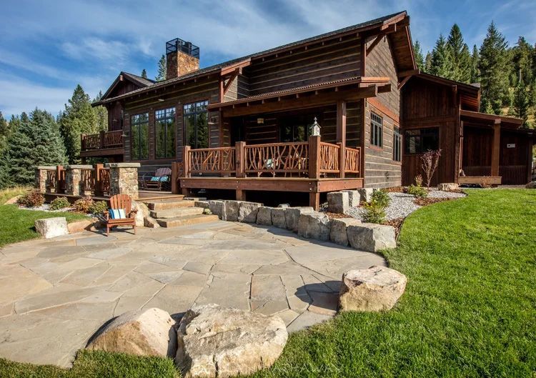A rustic log cabin with a stone patio, wooden porch, and surrounding forest on a sunny day.