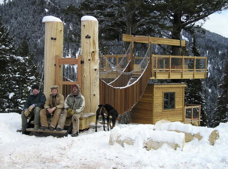 Three people sit on a wooden bench in front of a treehouse-style structure with a suspension bridge, set in snowy woods.