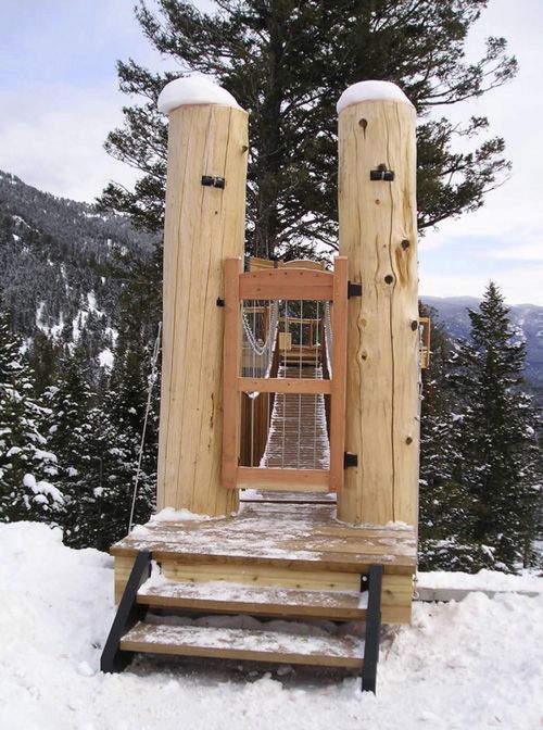 Wooden stairs lead to a gated entrance between two tall logs, opening onto a suspension bridge in a snowy, wooded setting.