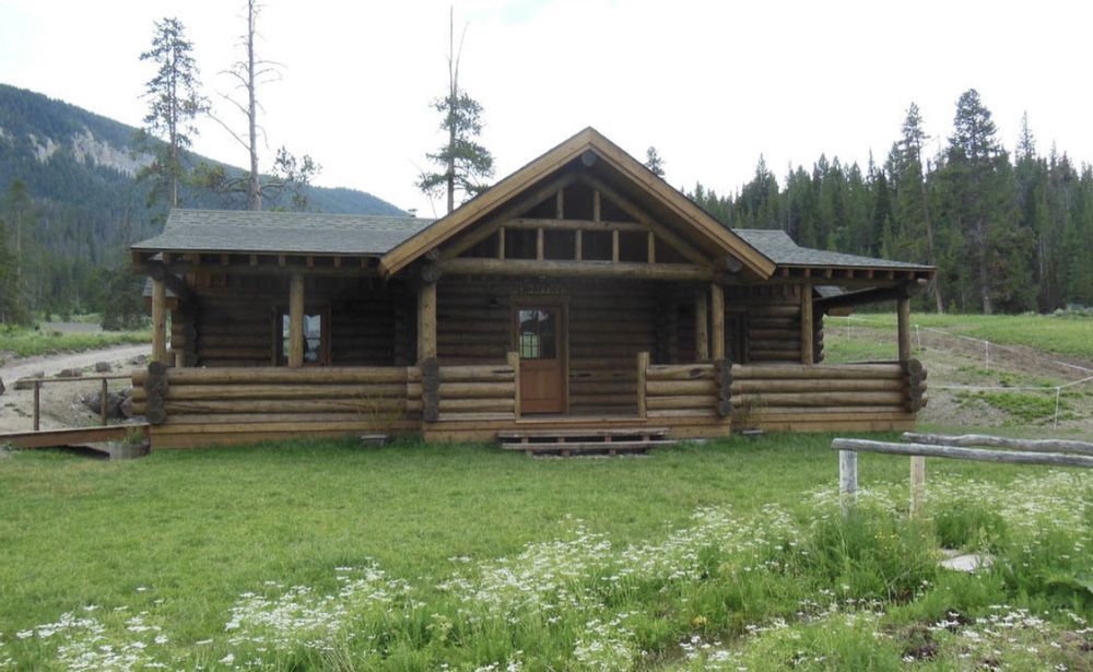 A rustic brown log cabin with a covered front porch, situated in a green meadow with a forest and mountain backdrop.