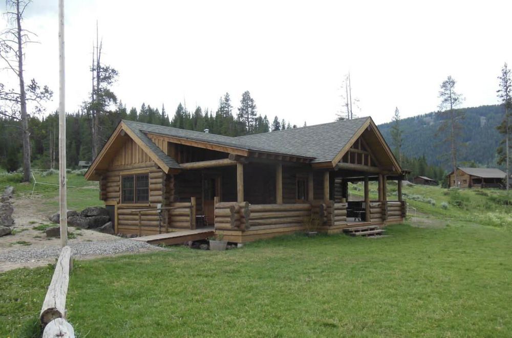 A rustic log cabin with a covered porch sits in a green, grassy field against a backdrop of trees and a mountain.