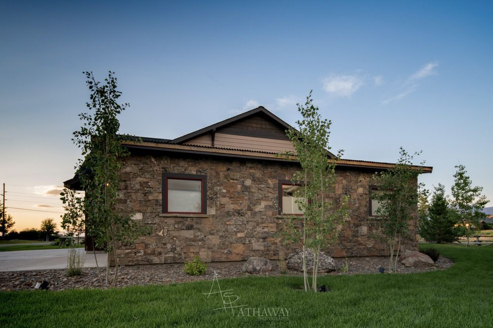 A stone house with brown horizontal siding under a gabled roof, surrounded by small trees and grass at sunset.