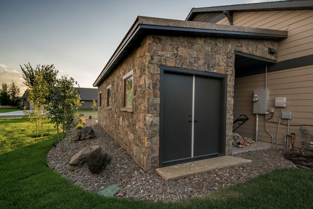 A stone-clad storage shed with dark double doors, set beside a residential house with tan siding and a gravel landscape.
