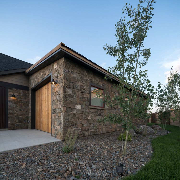 A side view of a stone garage with a wooden door and corrugated roof, set next to small trees, rocks, and grass at dusk.