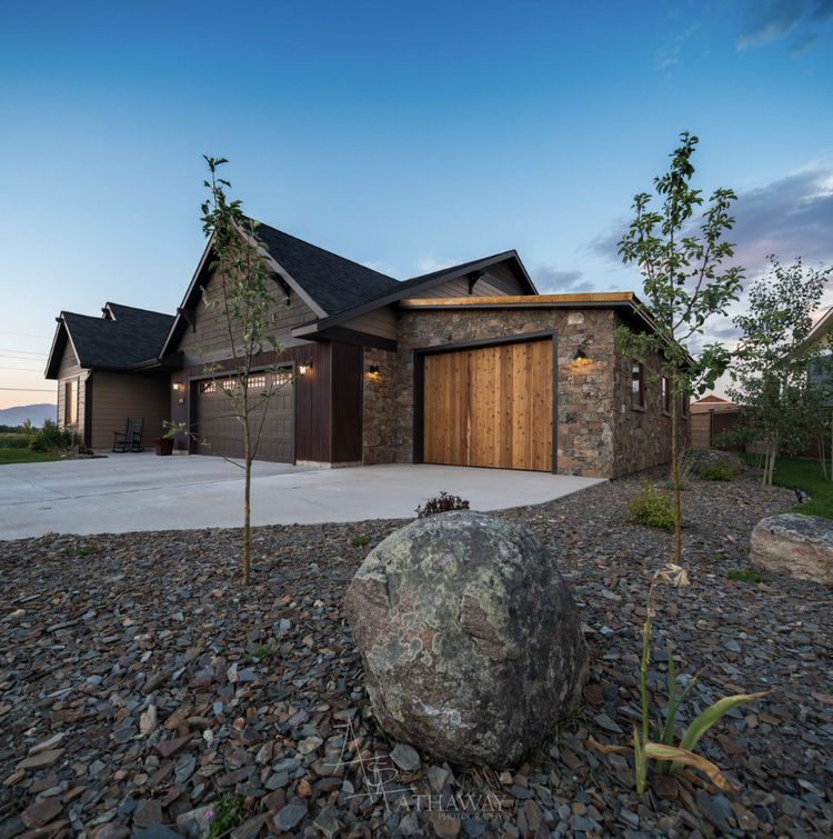 A dark-toned suburban house at twilight with a stone-accented garage, concrete driveway, and rock-covered front yard.