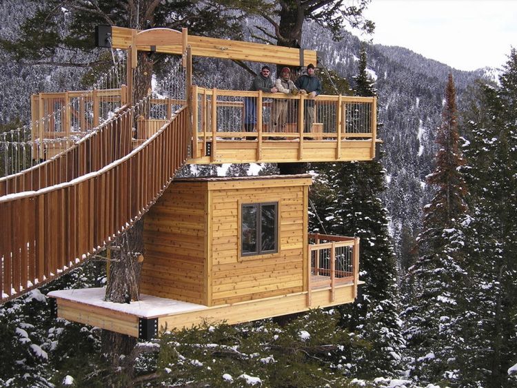 A wooden treehouse with a viewing deck connected by a bridge, nestled in snowy, forested mountains.