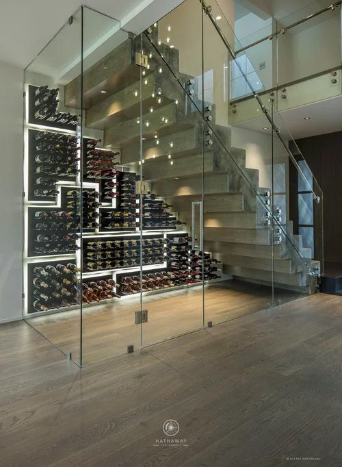 A glass-enclosed wine cellar built under a modern wooden staircase in a bright, contemporary home interior.