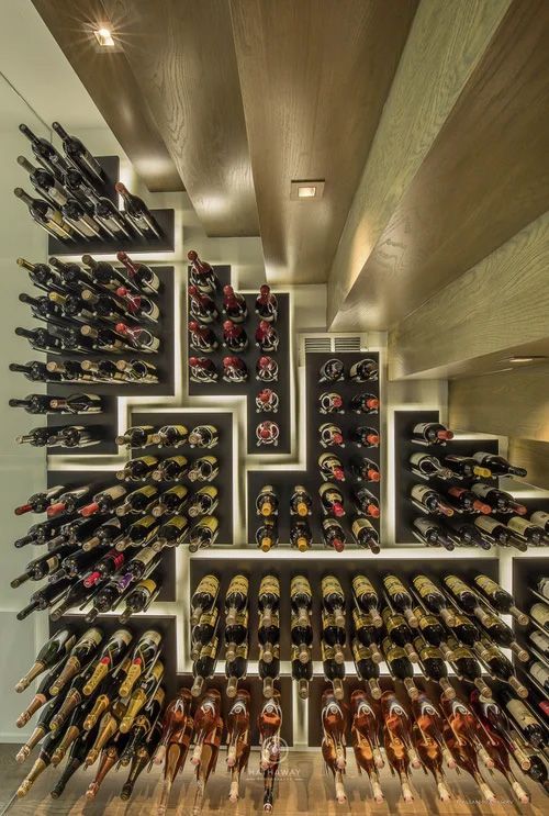 A modern, backlit geometric wine rack features rows of wine bottles stored on tiered, dark wooden shelves.