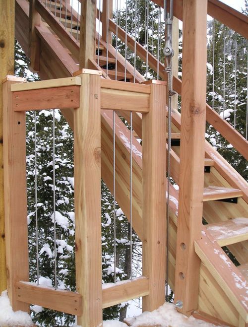 Outdoor wooden staircase with cable railing, surrounded by snow-covered trees.