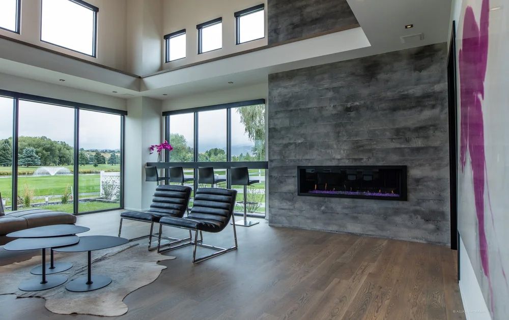 A bright, modern living room with large windows, leather chairs, wood floors, and a stone wall featuring a fireplace.