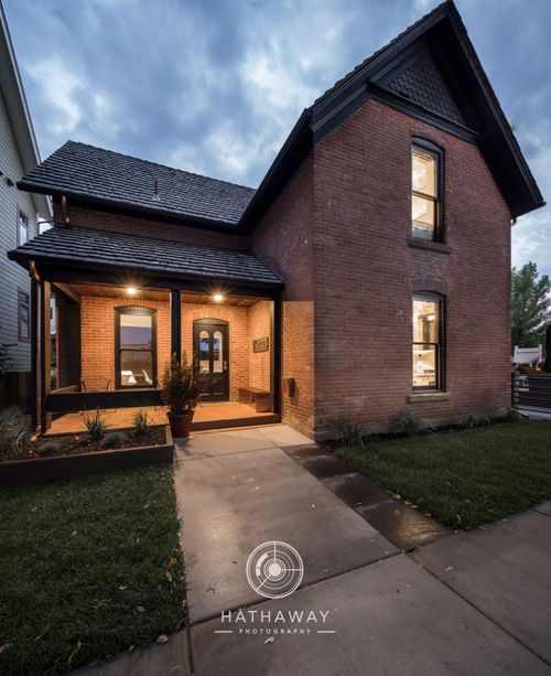 A two-story brick home at dusk with a dark-trimmed front porch and illuminated windows facing a paved walkway.
