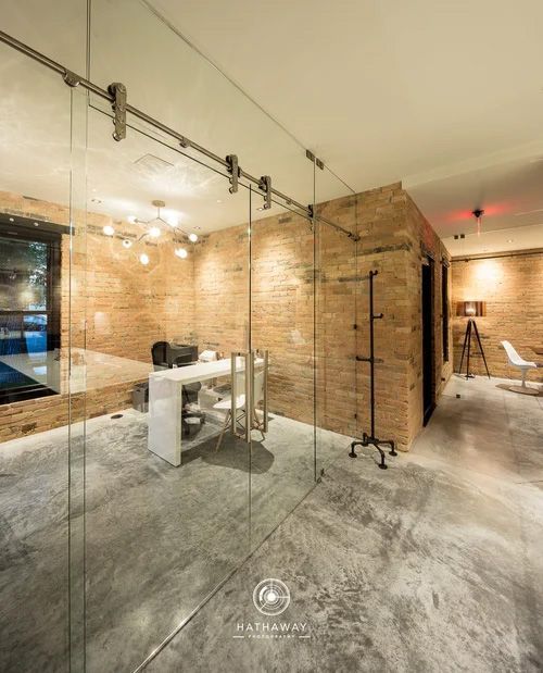 A glass-walled office with a white desk and modern lighting, set against a stone brick wall in a contemporary interior.