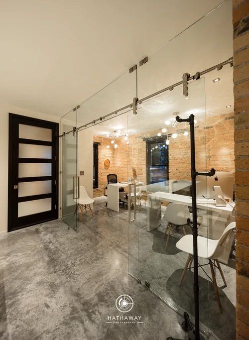 A modern office space with a glass sliding door, polished concrete floors, brick walls, and white desks and chairs.