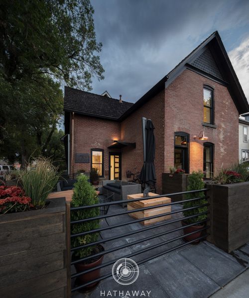 An evening view of a brick house featuring a modern, fenced patio with planters, outdoor seating, and warm lighting.